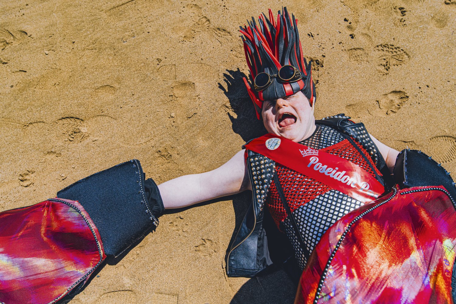 Eine Person in einem schwarz-roten Krabbenkostüm im Punk-Stil liegt auf dem Rücken im Sand. Die Person trägt eine Netz- und Nietenweste, eine rote Schärpe mit der Aufschrift "Poseidon", eine stachelige Kopfbedeckung sowie große Scherenhände und hat den Mu