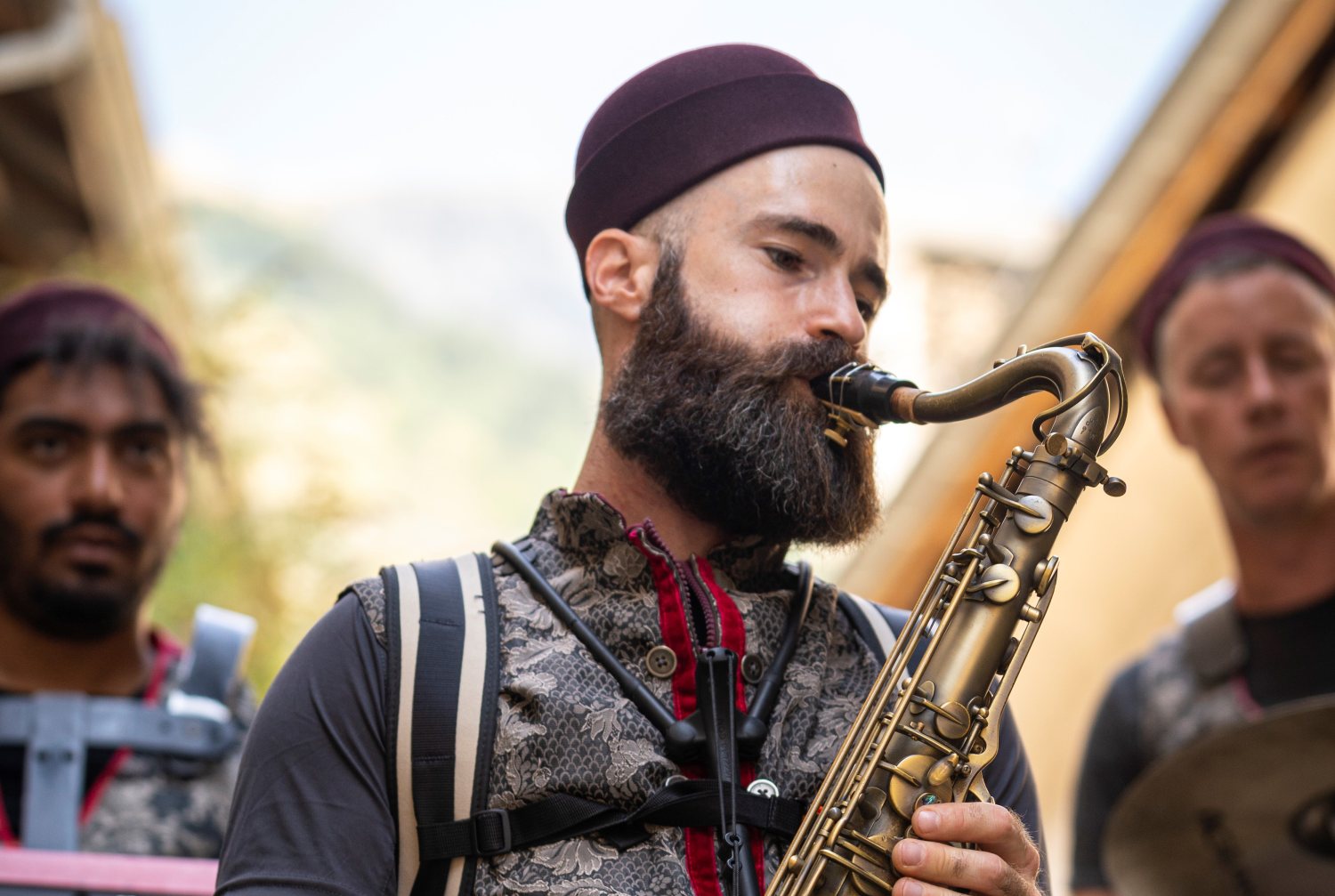 Nahaufnahme einer Person mit Vollbart beim Saxophonspielen. Die Person trägt eine gemusterte Weste mit roten Akzenten und eine weinrote Kappe. Im unscharfen Hintergrund sind zwei weitere Personen zu erkennen.