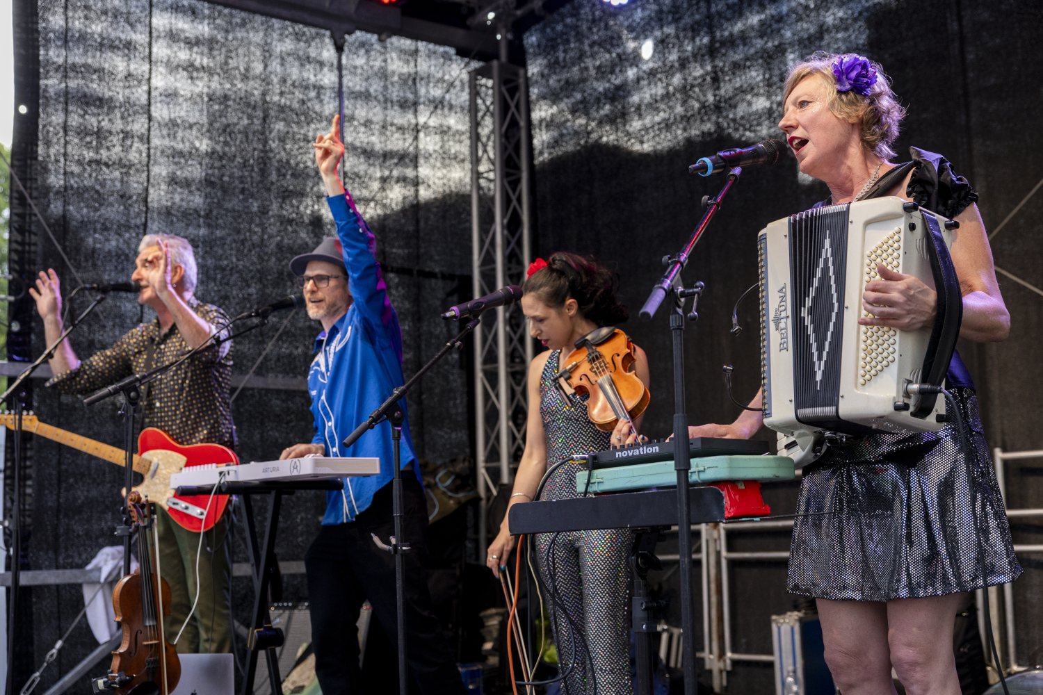 Die vierköpfige Band in Aktion auf der Bühne. Rechts singt die Person am Akkordeon, daneben bedient eine Person mit Geige in der Hand einen kleinen Synthesizer. Die Person am Keyboard und die Person an der E-Gitarre auf der linken Seite haben schwungvoll 