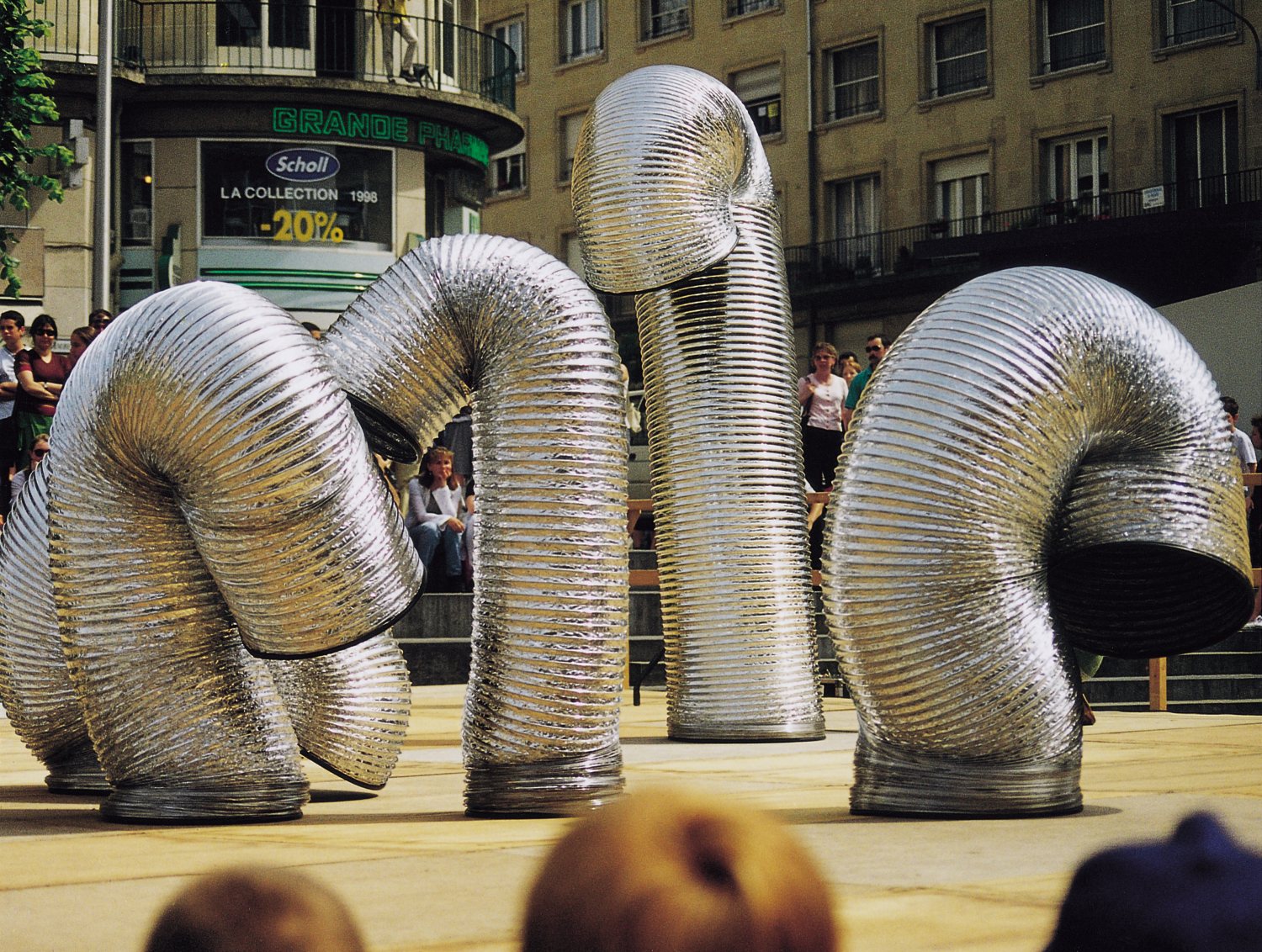 Mehrere riesige, flexible, silberne Röhren stehen und winden sich auf einem Platz und wirken wie überlebensgroße, abstrakte Figuren. Im Hintergrund steht eine Gruppe von Zuschauenden auf einer Treppe vor mehrstöckigen Gebäuden und beobachtet die Darbietun