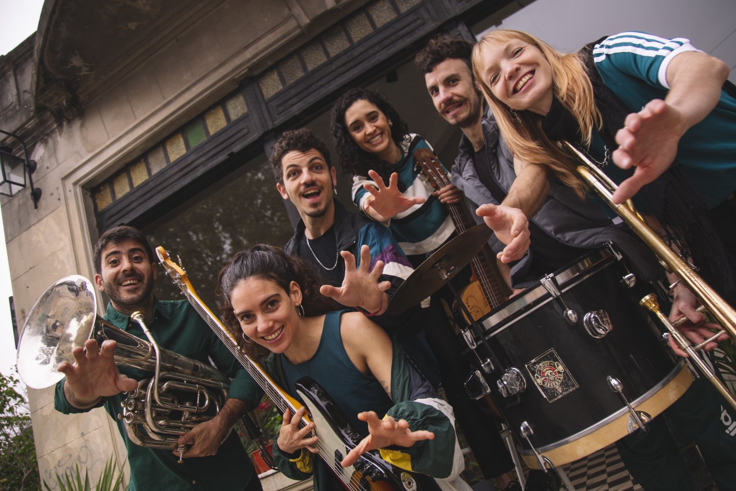 Eine Gruppe von sechs Musiker*innen posiert lachend vor einem Gebäude. Sie haben Instrumente wie Tuba, E-Bass, Akustikgitarre, Posaune sowie ein Schlagzeug bei sich und strecken jeweils eine Hand dynamisch in Richtung der Kamera.