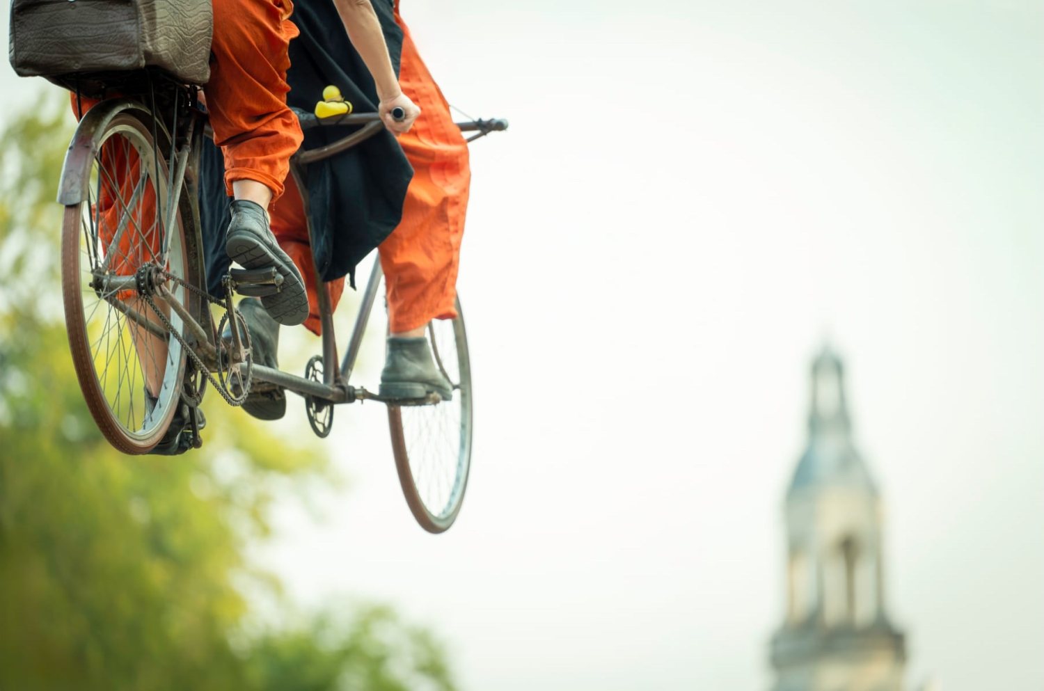 Zwei Personen in weiten, orangen Hosen fahren auf einem ungewöhnlichen Fahrrad, das scheinbar durch die Luft schwebt. Im unscharfen Hintergrund ist ein Turm zu erkennen.
