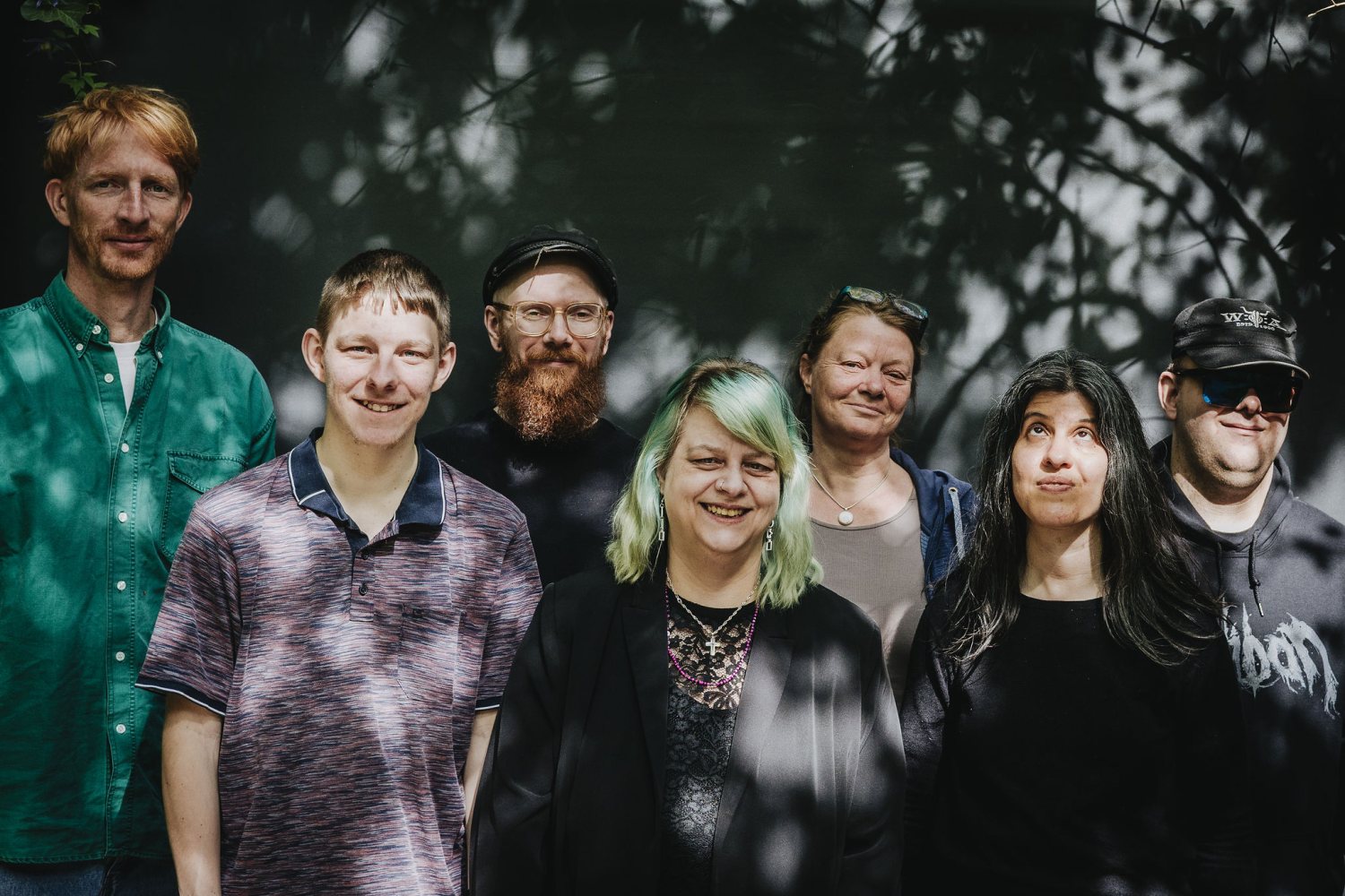 Ein Gruppenfoto von sieben Personen, die dicht beieinander im Freien vor einem dunklen, blättrigen Hintergrund stehen. Sonnenlicht fällt durch das Blätterdach fleckig auf die Gesichter der Gruppe.