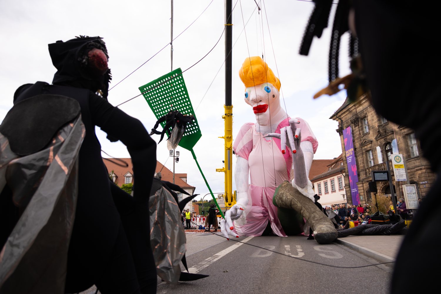 Eine überlebensgroße Puppe im rosa Kleid hockt auf der Straße. Im Vordergrund stehen in Schwarz gekleidete Akteur*innen mit transparenten Fliegenflügeln, während eine gigantische grüne Fliegenklatsche an Seilen über ihnen in der Luft hängt.
